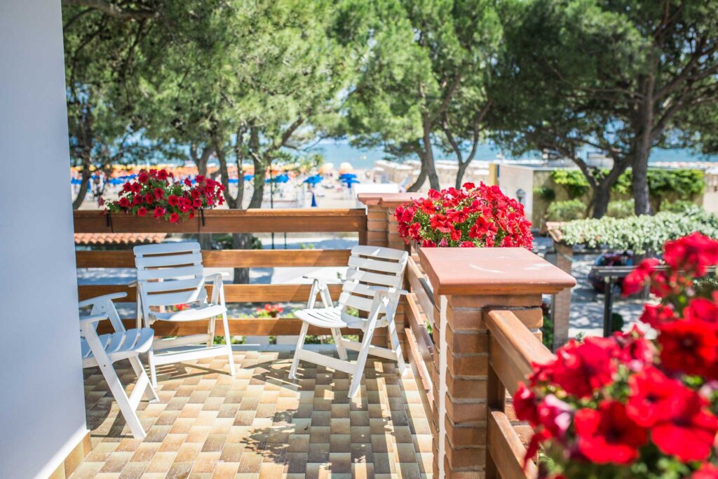 Terrazza con sedie bianche e fiori rossi, affacciata su una spiaggia alberata.