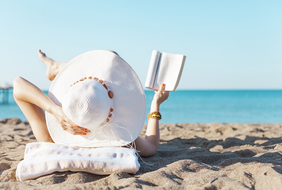 Donna su una spiaggia che legge un libro indossando un grande cappello bianco.